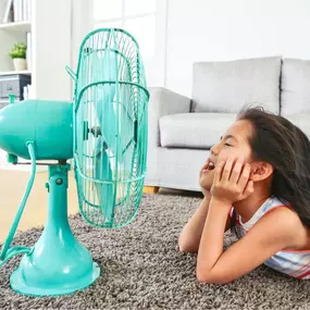 A young girl sits in front of a small desk fan, enjoying the cool breeze as she tries to stay comfortable on a hot day. Her hair blows gently in the airflow, and her relaxed expression shows relief from the heat—suggesting the absence of central air conditioning or the need for better indoor cooling. This image highlights the importance of reliable A/C, energy-efficient HVAC systems, and summertime home comfort.