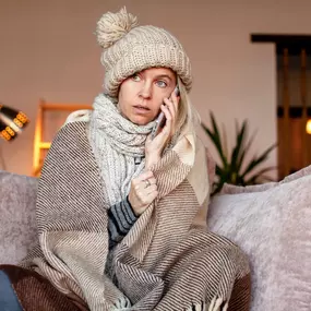 A woman bundled in winter clothing sits indoors, visibly cold as she makes a phone call—likely reaching out for emergency heating repair. Wearing a scarf, hat, and heavy jacket, she appears uncomfortable due to a broken furnace or malfunctioning heating system. This image highlights the urgency of reliable HVAC service, the importance of routine maintenance, and the need for fast, professional heating repairs during cold weather.
