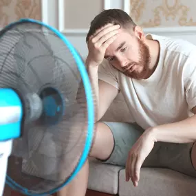 A man sits indoors in front of a small electric fan, wiping sweat from his forehead as he tries to stay cool during a heatwave. His expression shows discomfort, suggesting that his air conditioning may be broken or insufficient. This image highlights the struggle of staying cool without proper HVAC, emphasizing the need for reliable air conditioning service, energy-efficient upgrades, and regular A/C maintenance.