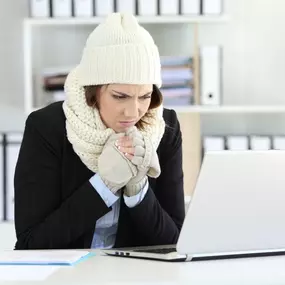 A woman bundled up in a hat and jacket, sits in her chilly home trying to stay warm. She appears uncomfortable, layered in winter clothes, and clearly affected by the cold indoor temperature—possibly due to a broken heater or inefficient furnace. This image perfectly illustrates the importance of reliable home heating, timely HVAC maintenance, and professional furnace repair during the winter months. Great for use in heating service promotions or cold weather safety tips.