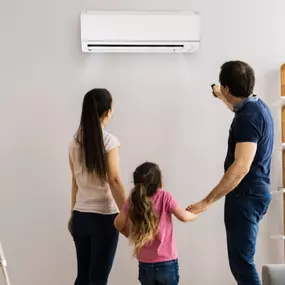 A smiling family stands together admiring their newly installed mini-split air conditioning unit. Mounted high on the wall inside their home, the sleek and quiet system provides efficient cooling without the need for ductwork. The parents and children look pleased with the comfort and energy savings the ductless system offers. This image highlights the benefits of modern HVAC technology, showcasing mini-split A/C installation for residential homes seeking zoned cooling, lower utility bills, and 