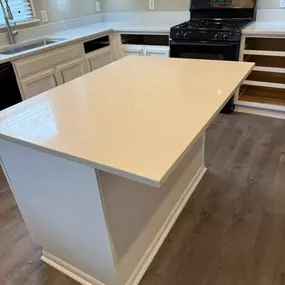 This image shows a kitchen in progress, featuring white cabinets and pristine white countertops. A central island with a matching white top takes prominence. A black stove is installed, and some cabinet spaces are still open, awaiting completion.
