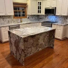 This image showcases a newly renovated kitchen with white cabinets and striking granite countertops and backsplash. A large, matching granite island with waterfall edges anchors the center of the room, complemented by warm hardwood floors.