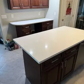 This image shows a kitchen or office nook with dark wood cabinetry and pristine white countertops. A central island provides additional workspace, and a calendar hangs on a nearby white door, adding a touch of color.