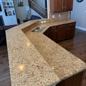 This image showcases a kitchen with speckled beige granite countertops forming an L-shape, incorporating a double sink. Dark wood cabinets complement the rich wood flooring, with a staircase and living area visible in the background.