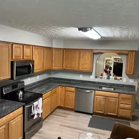 This image displays a kitchen with warm-toned wooden cabinets and dark, speckled countertops. Stainless steel appliances, including a microwave, stove, and dishwasher, are integrated into the design. Light-colored wood flooring completes the space.