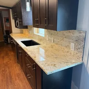 This image showcases a kitchen with sleek dark wood cabinets complemented by light-speckled granite countertops and a matching backsplash. A black sink is integrated, and a modern pendant light hangs above the rich wood flooring.