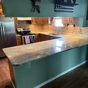 This image features a kitchen with light wood cabinets and brown tiled backsplashes. Speckled light granite countertops with an integrated sink and an electric stove are visible. A patriotic-themed shelf and star decor adorn the sage green walls.