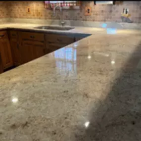 This image offers a close-up view of a polished granite countertop, displaying a speckled pattern in shades of beige and brown, reflecting light. In the background, part of a kitchen with light wood cabinets and a tiled backsplash is visible.