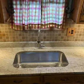 This image focuses on a kitchen sink area, featuring a shiny stainless steel sink set into a light, speckled granite countertop. Above, light wood cabinets are visible, along with a brown tiled backsplash and a plaid curtain in the window.