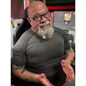 In this dynamic and approachable portrait, Connor MacIvor—known as “Connor with Honor”—flashes a charismatic smile while seated in his content creation studio. His muscular frame, trimmed beard, and confident body language reflect the discipline behind his 135-pound weight loss transformation and ongoing commitment to health, leadership, and authenticity. Captured mid-conversation with hands open in a welcoming gesture, the image shows Connor's natural energy and warmth—qualities that make him a