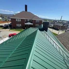 A newly installed green metal roof viewed from above, showcasing clean lines and precise construction. Surrounding neighborhood homes and buildings are visible under a bright, clear blue sky.