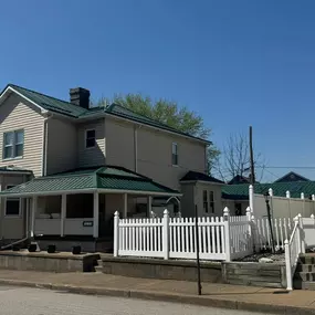 A two-story beige home with green metal roofing and a covered porch, surrounded by a white picket fence. The well-kept yard and neighboring homes sit beneath a clear blue sky.