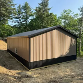 A newly built tan and black metal structure with a sloped roof stands on a dirt clearing, surrounded by tall pine trees and dense forest under a clear blue sky.