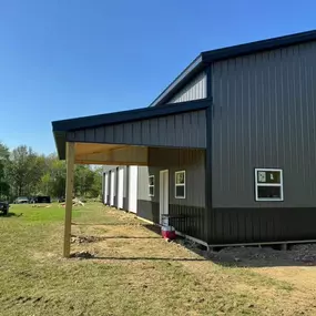Modern metal building with gray vertical siding, multiple white-trimmed windows, and a covered entryway under construction. The structure sits on a grassy plot surrounded by trees under a clear blue sky.