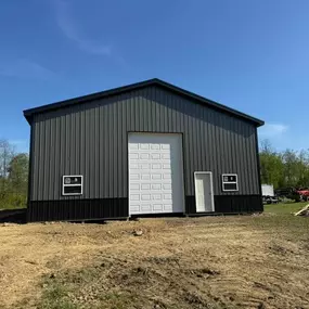 A large black metal building with a white garage door and entry door, featuring two windows. Surrounded by grass and light construction materials under a clear blue sky in a rural area.