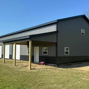 A large dark gray metal building with multiple white garage doors, a small porch area with wooden posts, and several windows. Surrounded by grass and trees, under a bright blue sky.