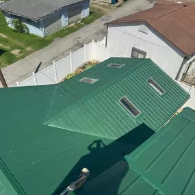 Aerial view of a green metal roof with multiple ridges and skylights, surrounded by a white privacy fence and adjacent buildings. Clean, precise installation highlights professional craftsmanship and roofing detail.