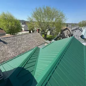 View from a rooftop showing a newly installed green metal roof adjacent to an older shingle roof. A neighborhood of homes and trees stretches out beneath a clear blue sky.
