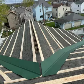 Roof installation underway with wood battens installed over old shingles, preparing for metal roofing. A green valley panel is placed, and surrounding homes and trees frame the residential construction site.
