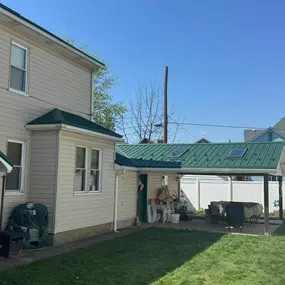 A well-maintained beige house with a green metal roof, skylights, and a covered patio area. The backyard features a grassy lawn, white fence, and patio furniture under a shaded structure.