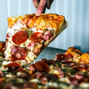 Hand pulling a loaded slice of pizza from a fresh whole pizza, topped with pepperoni, ham, and melted cheese.