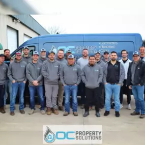 A team from OC Property Solutions poses in front of their service van, representing their full-service restoration, plumbing, electrical, and HVAC company.