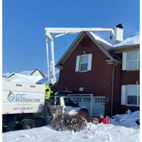 Crew members are performing winter tree services using a bucket truck outside a snow-covered residential home.