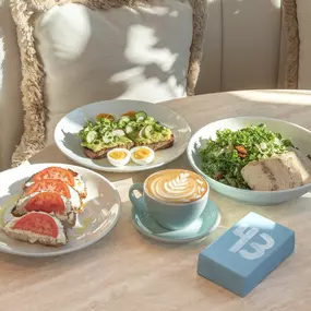 All day brunch table spread with tomato bagel, avocado smashtoast with soft-boiled eggs, kale caesar salad with chicken andalmond latte.