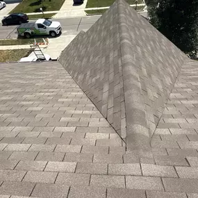 A top-down view of a residential roof covered in light brown asphalt shingles. The image highlights a sharp, prominent ridge line and a white work truck in the street below.