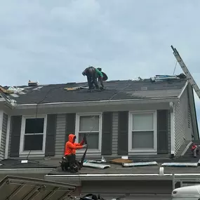 A view of three roofers working on a residential home with gray siding. One is on the top roof, and two are on the lower roof, which is in the process of being torn off.