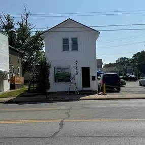 A tall, narrow, two-story white building with a black door and large window. A ladder leans against the side, a sign is in the window, and a car is parked nearby.