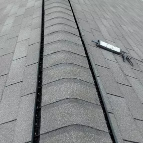 A close-up, high-angle view of a gray shingle roof, showing a newly installed ridge vent running along the peak. A caulking gun rests on the shingles nearby.