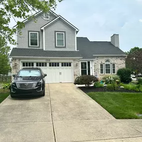 A suburban two-story house with a two-car garage, gray siding, and light-colored brick. A black SUV is parked in the driveway, and a well-kept lawn and garden are visible.