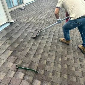 A close-up of a worker in jeans and work boots using a long-handled shingle remover to tear off old brown shingles from a roof, with a pry bar lying nearby.
