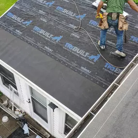 A top-down view of a worker on a residential roof installing synthetic underlayment. The roof is partially covered with the black material, and a red shed is visible in the background.
