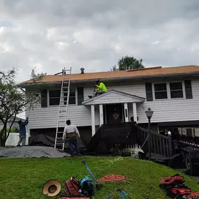 A residential home with three workers on a roof, in the process of replacing the shingles. Tarps are on the lawn, and a large black trailer is parked in the driveway.