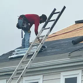 A side view of a roofer in a red shirt and jeans on a roof, preparing to lay down black roofing underlayment. A ladder and sacks of materials are nearby.