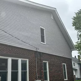 A residential building's exterior, showing the transition from brown brick on the first floor to gray vinyl siding on the upper story and gable end. A tall window is visible.