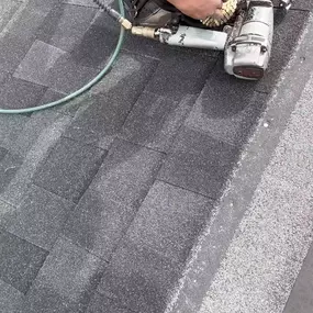 This close-up aerial shot captures a person wearing a maroon long-sleeve shirt and gloves, using an air-powered nail gun to secure dark gray asphalt shingles onto a roof, showing the precise process of roofing installation.