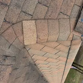 This image captures an overhead view of a weathered brown shingle roof, showing a valley where two roof sections meet. The shingles display signs of wear and tear, with some discoloration and missing granules, under an overcast sky.