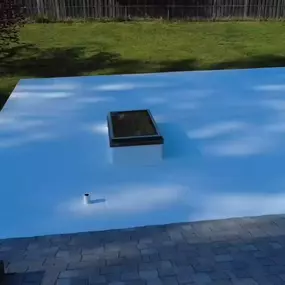 This image presents a large, newly installed white flat roof with three skylights, reflecting sunlight. Below, part of an existing gray shingle roof is visible, and in the background, a green lawn and trees under a clear sky.
