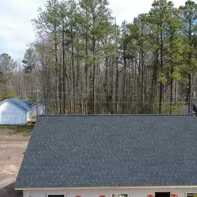 This image captures an aerial view of a new home construction with a dark shingle roof, set against a backdrop of tall, green pine trees. Other houses and a road are visible in the distance under a blue sky.