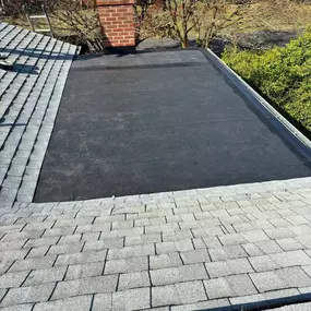 This image shows a roof with both gray shingle sections and a large, flat black rubberized area, suggesting a repair or combination roofing. A brick chimney stands tall, and trees with red leaves are visible in the background.