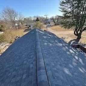 This image captures an overhead view of a newly installed dark gray shingle roof, extending towards the horizon under a clear blue sky. In the background, scattered trees and residential houses complete the suburban landscape.