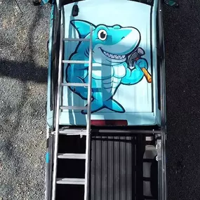 This overhead view of a light blue pickup truck shows a vibrant cartoon shark holding a hammer painted on its roof, emphasizing the 