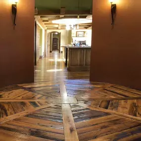The image showcases a distinctive starburst-patterned hardwood floor, possibly by Enmar or Richard Marshall Flooring, leading into a warmly lit home interior. The rustic wood contrasts with the elegant wall sconces and arched doorway.