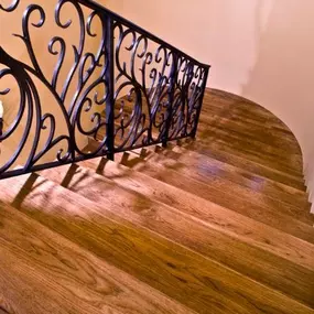 The image shows a gracefully curving wooden staircase, featuring rich brown treads that contrast elegantly with the dark, ornate metal railing. Soft lighting highlights the wood's texture and the railing's intricate design.