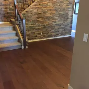 This image features a welcoming home interior with dark hardwood flooring and a staircase. A striking stone accent wall adds texture, complementing the stairs' carpeted treads and dark wood newel post and metal balusters.