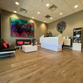 The image shows the modern interior of Flawless Faces Med Spa in Chandler, featuring a sleek white reception desk with blue lighting, a black sofa with pink pillows, and vibrant artwork on the walls.